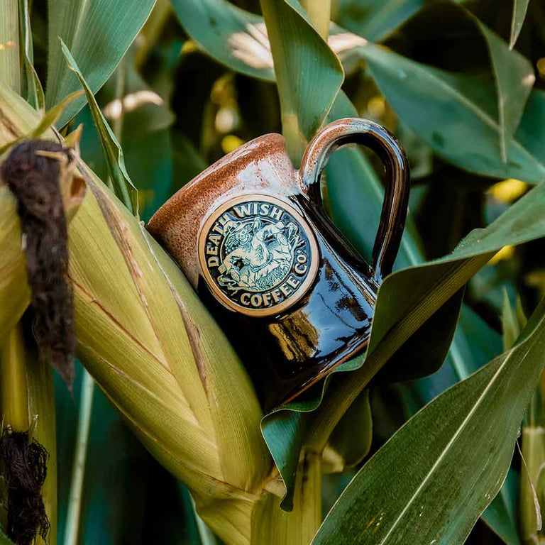 The Death Wish Coffee Handcrafted Scarecrow Mug resting on corn.